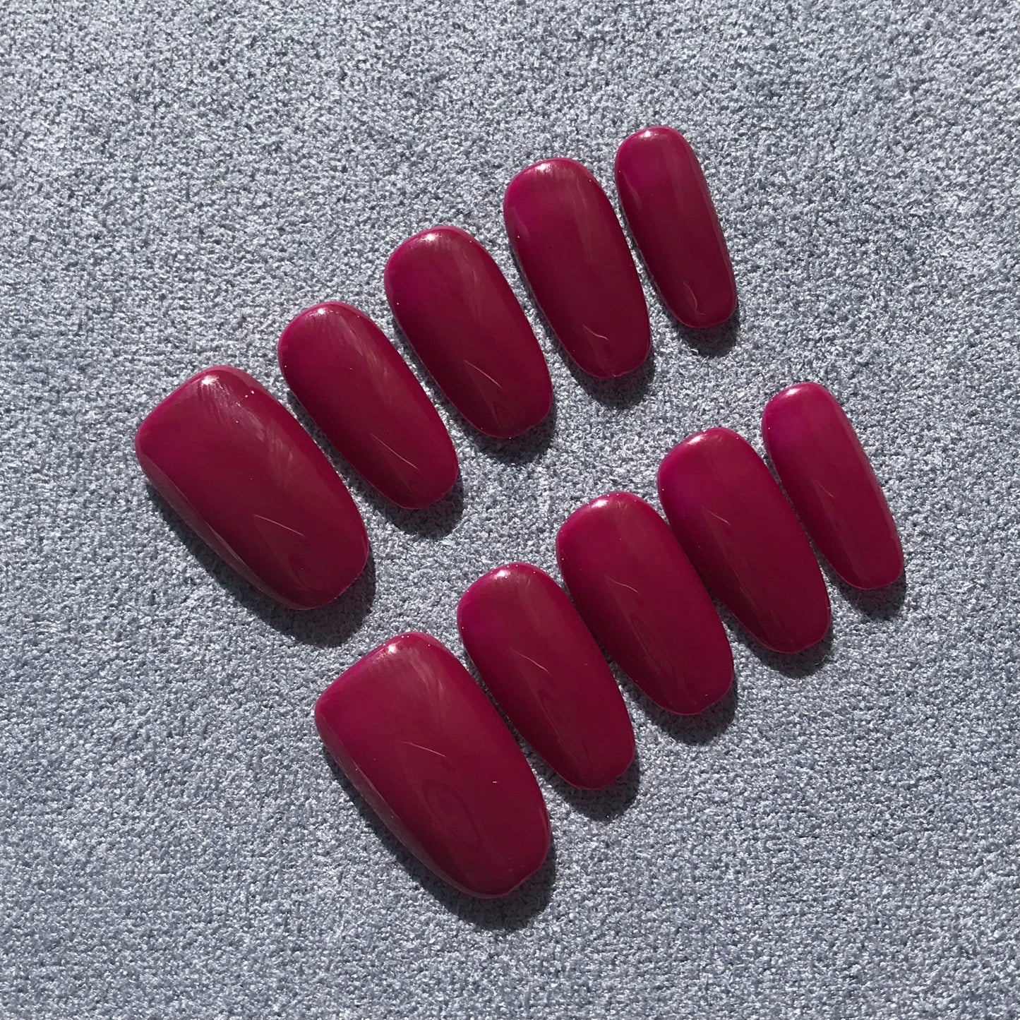 A set of ten press-on nails in a glossy deep magenta color, arranged on a textured gray fabric background.