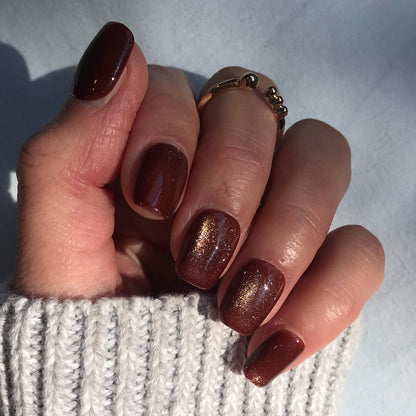 A close-up of a hand wearing press-on nails in deep metallic bronze with a subtle gold shimmer.