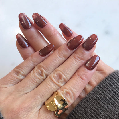 A close-up of hands modelling oval-shaped press-on nails in a glossy warm brown with fine gold glitter. 