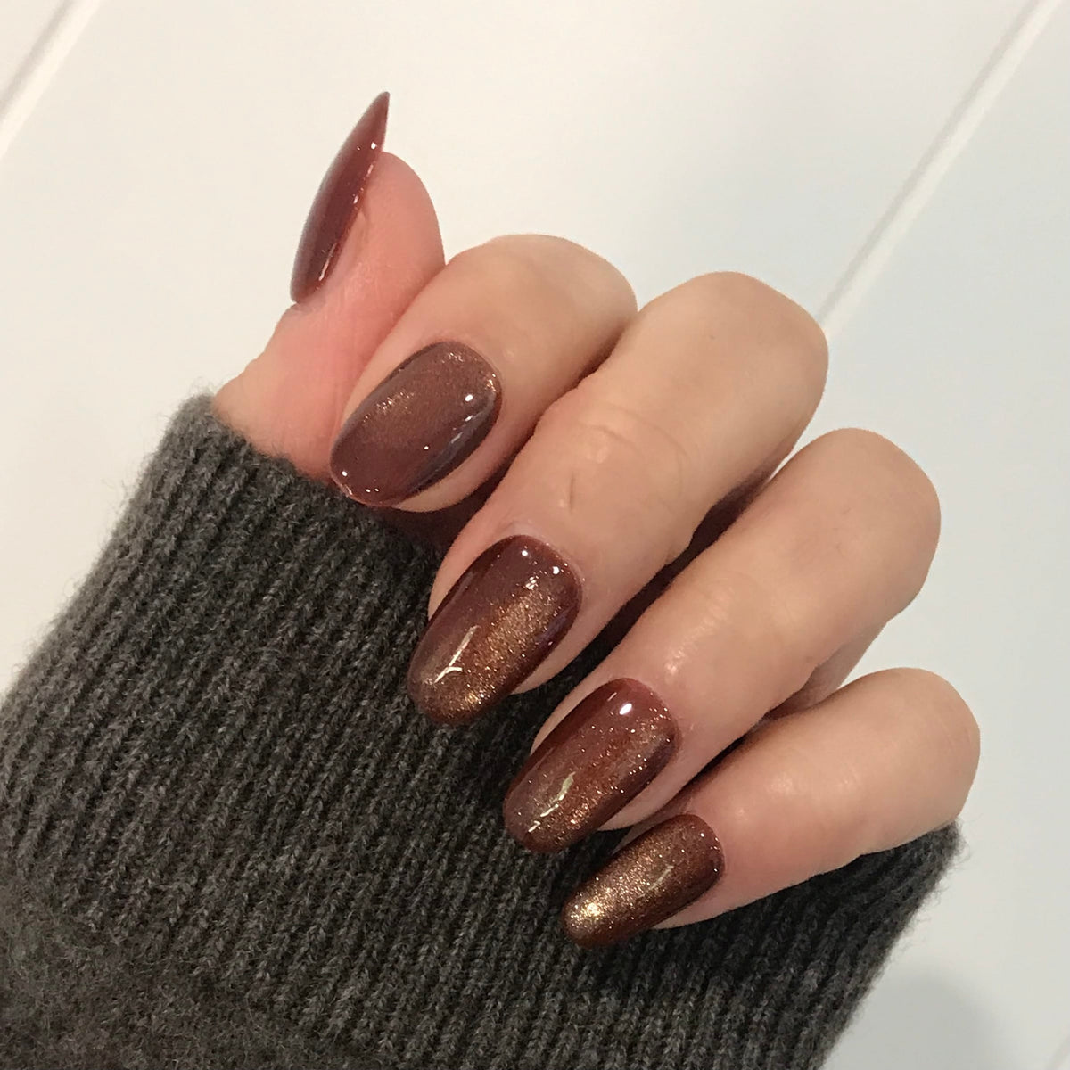 A close-up of a hand with oval-shaped press-on nails in glossy metallic bronze-brown.