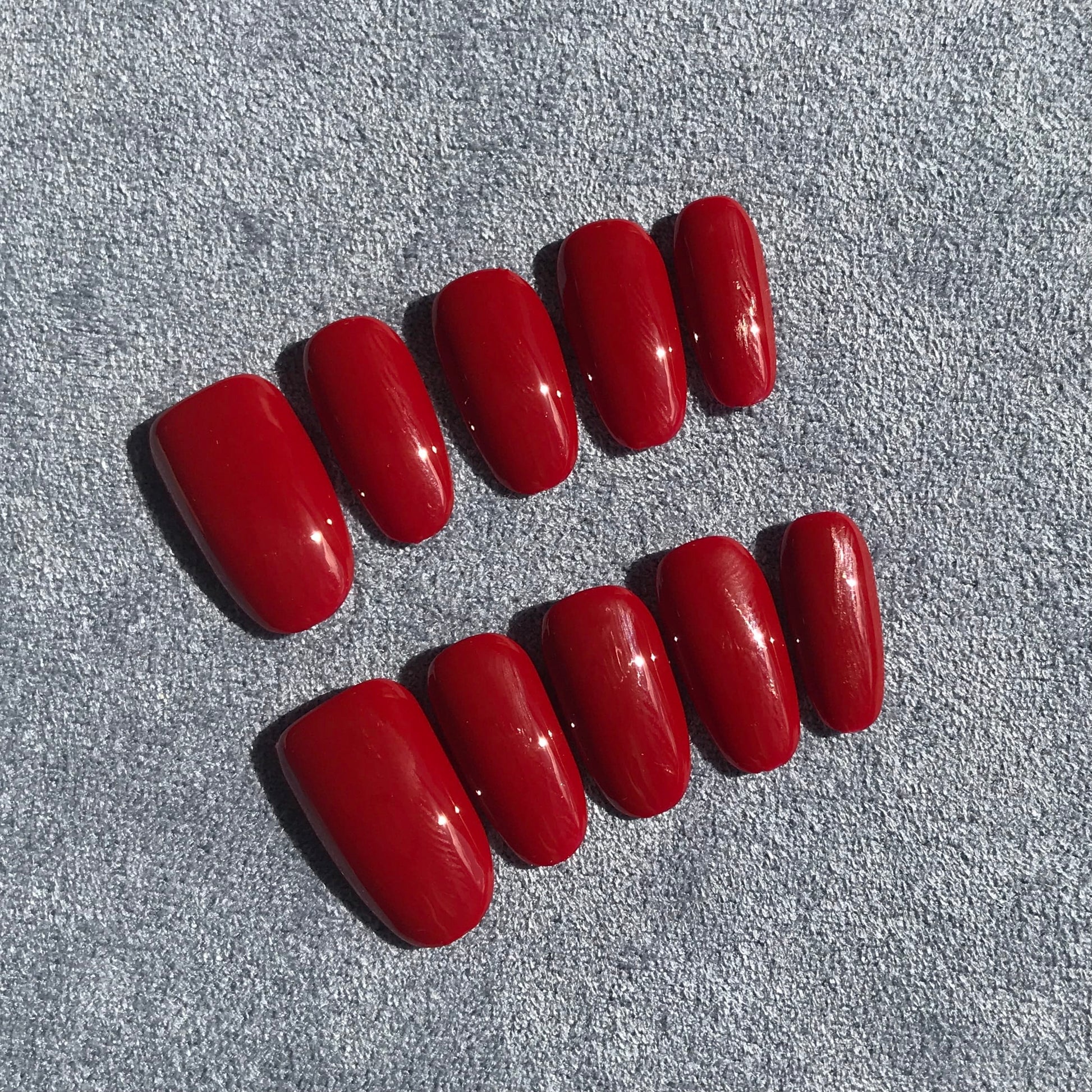 Ten red press-on nails with a glossy finish, on a textured gray background.