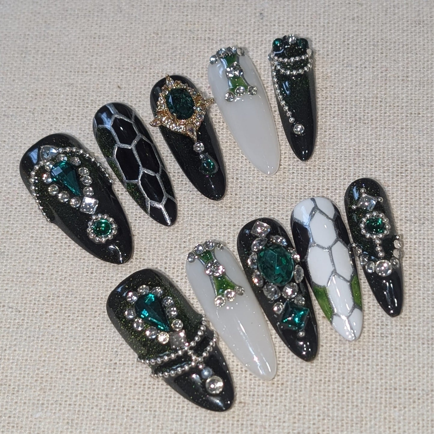 Handmade press-on nails featuring a mix of glossy black, white, and silver designs, embellished with rhinestones, green gemstones, and snake-scale patterns.