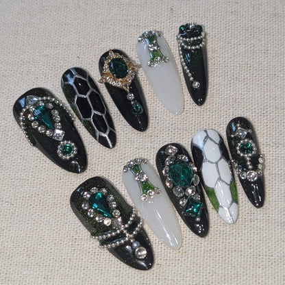 Handmade press-on nails featuring a mix of glossy black, white, and silver designs, embellished with rhinestones, green gemstones, and snake-scale patterns.