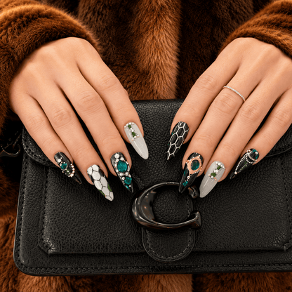 A close-up of handcrafted press-on nails decorated in green, black, white, and gold patterns with gem stones and silver beads. 