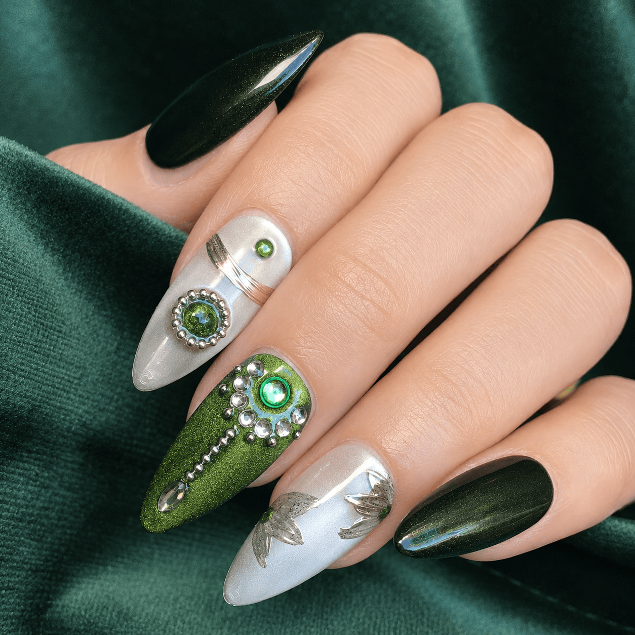 A close-up of a hand showcasing a set of long, almond-shaped press on nails with intricate nail art. The design features a mix of metallic olive green, shimmering dark green, and glossy white nails decorated with rhinestones, green gems, and silver leaf motifs.