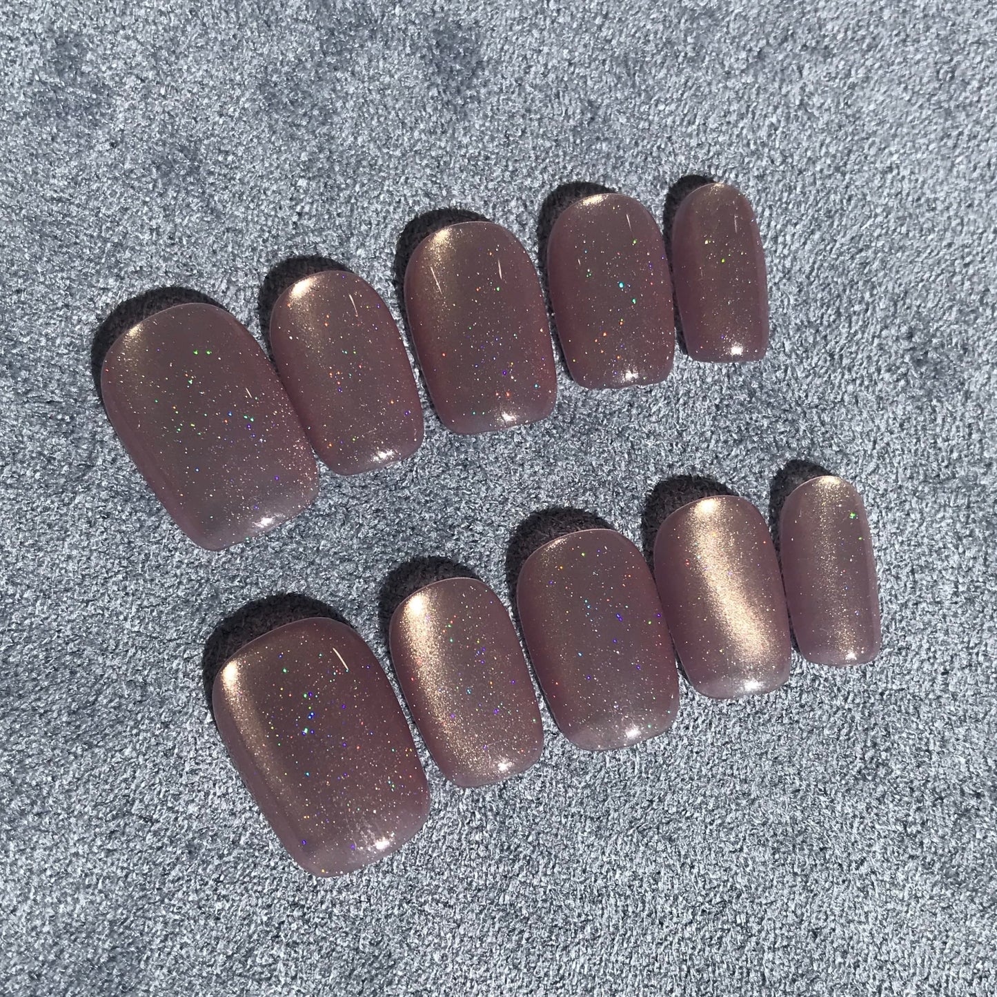 Ten press-on nails with a metallic lilac glitter finish, arranged in two rows on a textured background.
