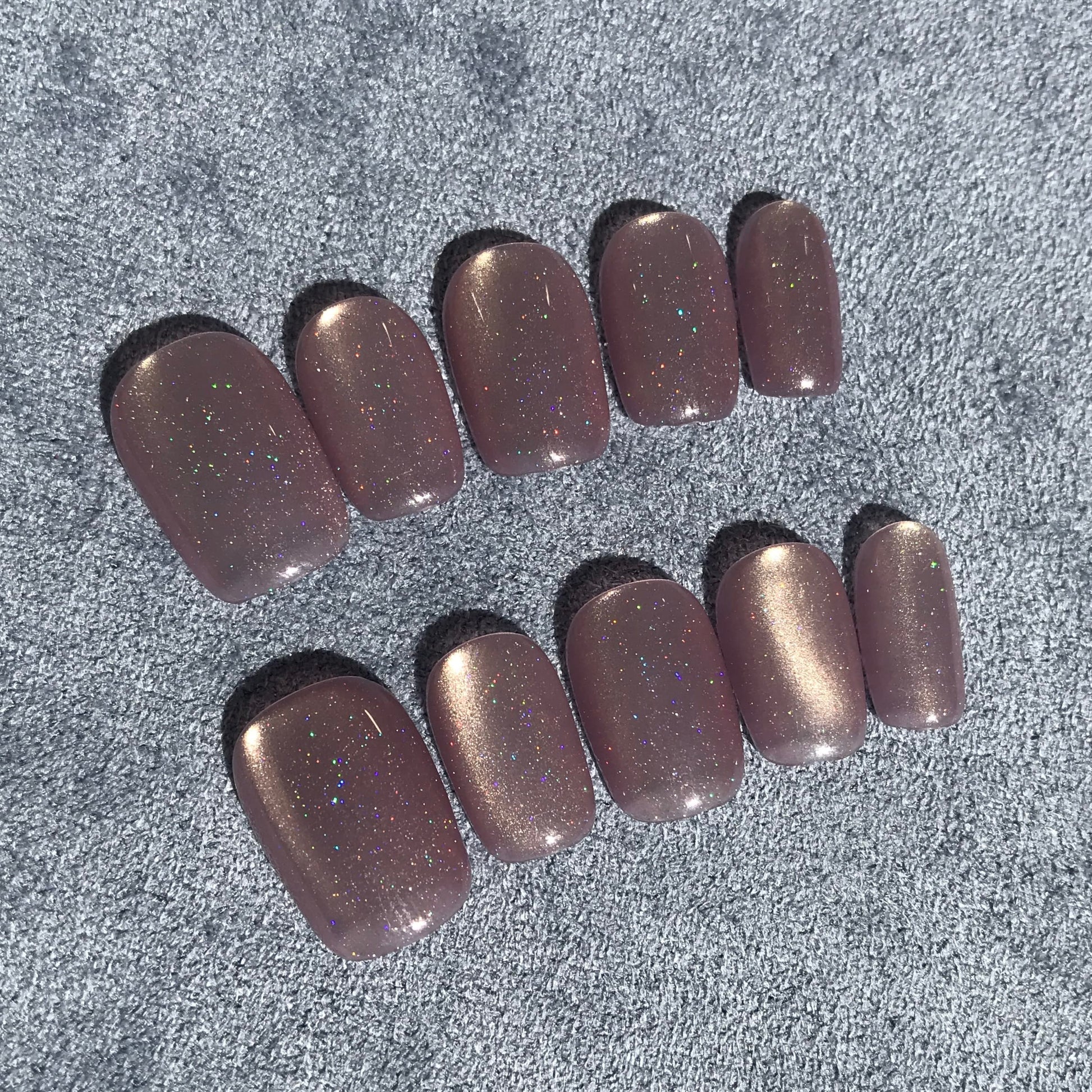 Ten press-on nails with a metallic lilac glitter finish, arranged in two rows on a textured background.