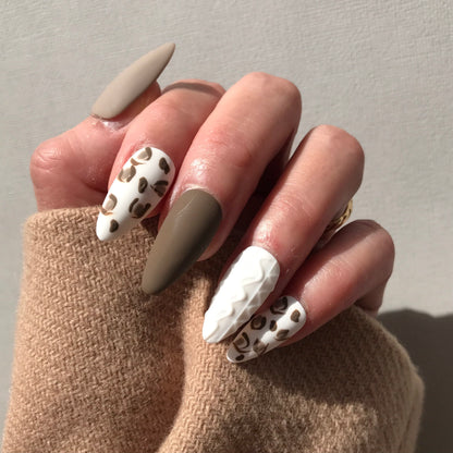 Hand with stylish press-on nails featuring beige, white, and leopard print designs.