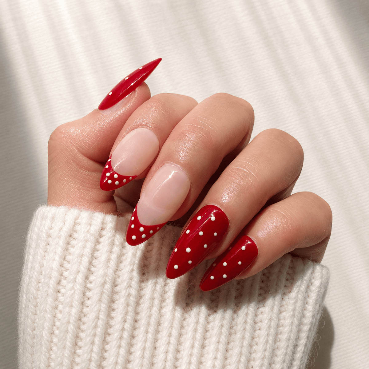 A close-up of a hand with almond-shaped press-on nails featuring a glossy red and white French tip with white polka dots.