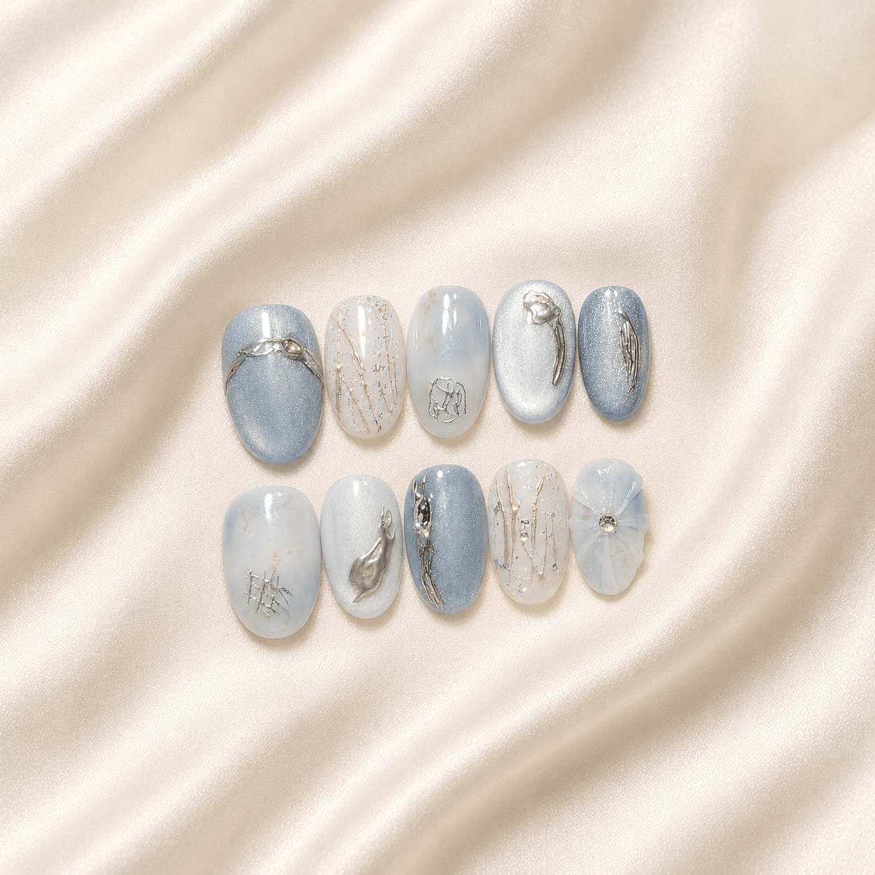 Ten press-on nails arranged in two rows on a soft cream satin fabric background. The nails feature a mix of light blue, white, and silver-gray designs with delicate metallic and abstract details.