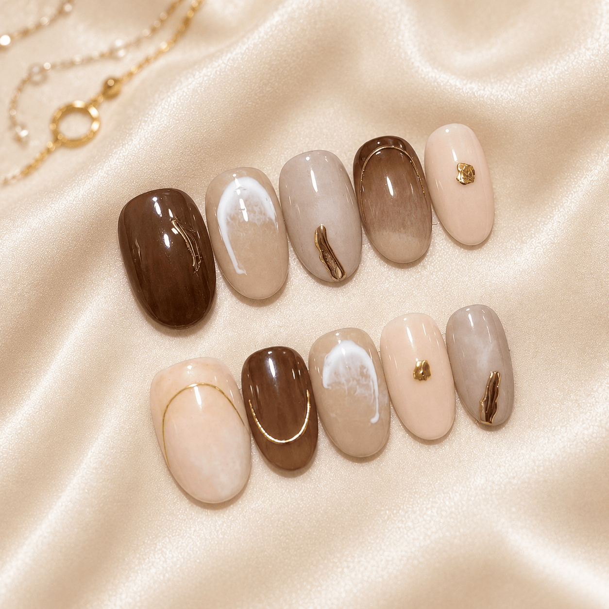 A set of 10 almond-shaped press-on nails displayed on a beige satin fabric background. The nails feature various designs including solid dark brown, glossy taupe, cream with cat-eye shimmer, and light pink with gold metallic accents.
