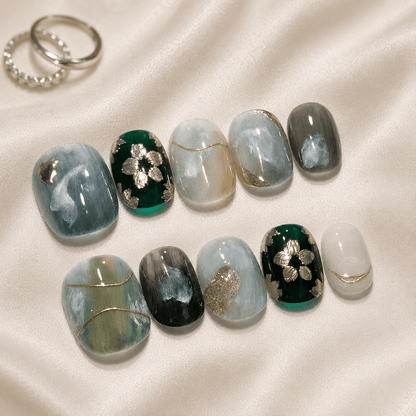 Decorative press-on nails arranged on a cream satin fabric background, The nails feature marbled blue, green, gray, black, and white patterns with metallic gold foil accents and floral motifs.