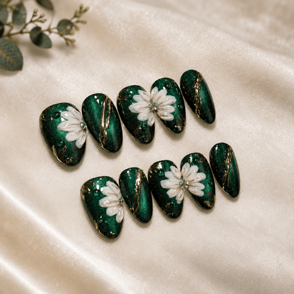 A set of ten almond-shaped press-on nails with a glossy dark green magnetic base, featuring hand-painted white daisy flowers and shimmering gold foil veins, arranged on a cream satin fabric background.